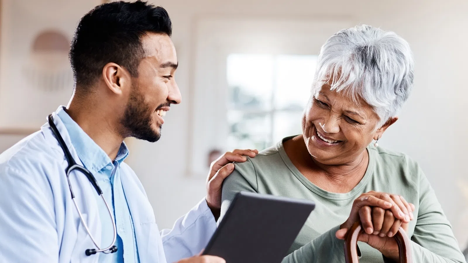 Smiling doctor with stethoscope showing tablet to elderly woman with cane in bright room
