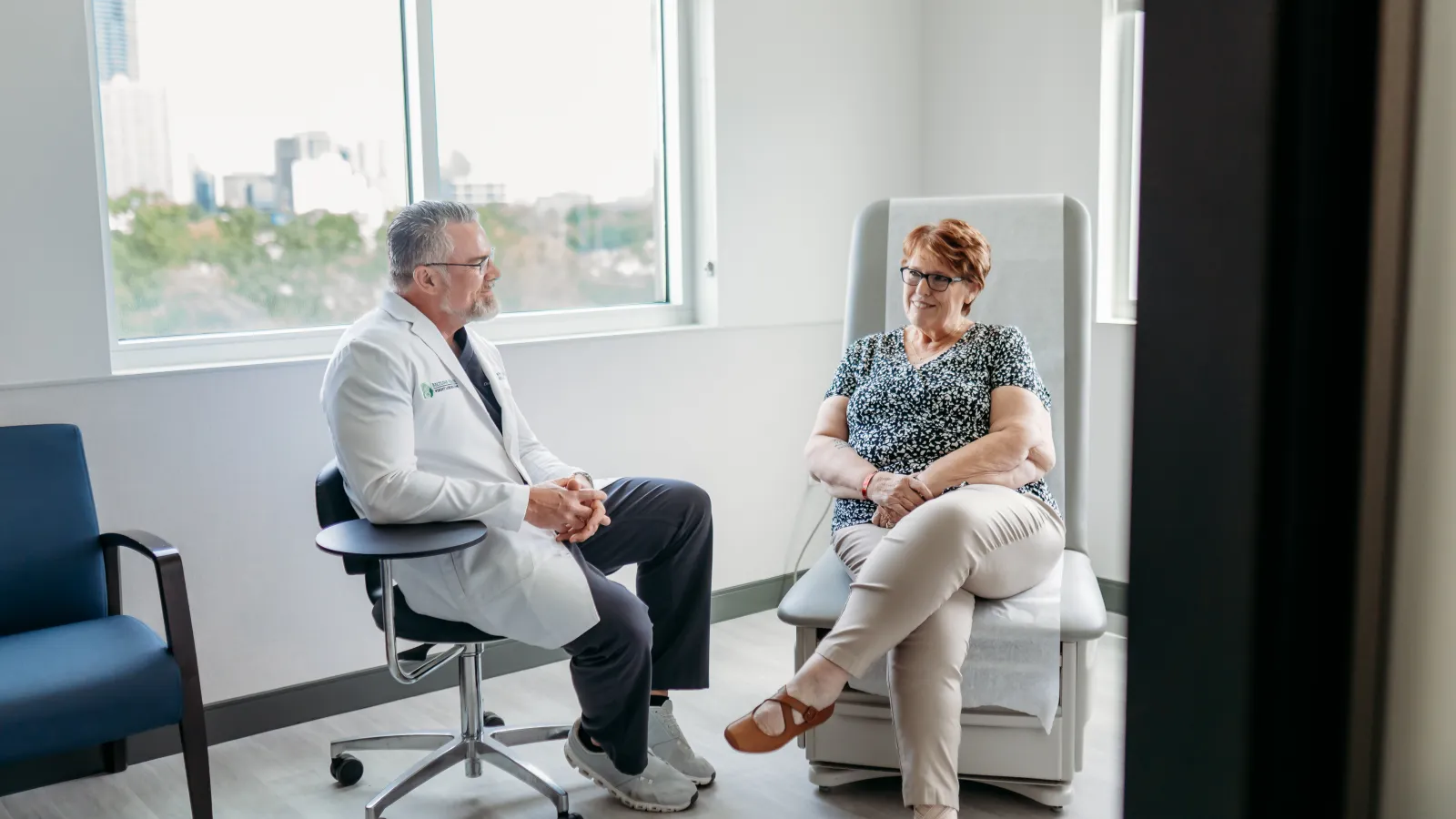 Doctor in white coat consulting with female patient in bright medical office with large windows.