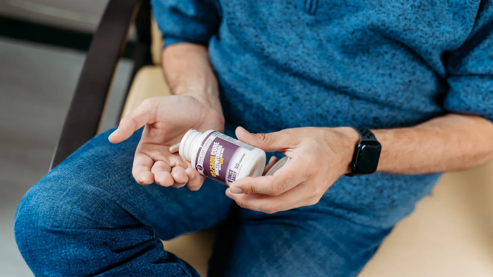 Man wearing blue sweater holding a bottle of capsules and placing pills in his hand while sitting on a chair.