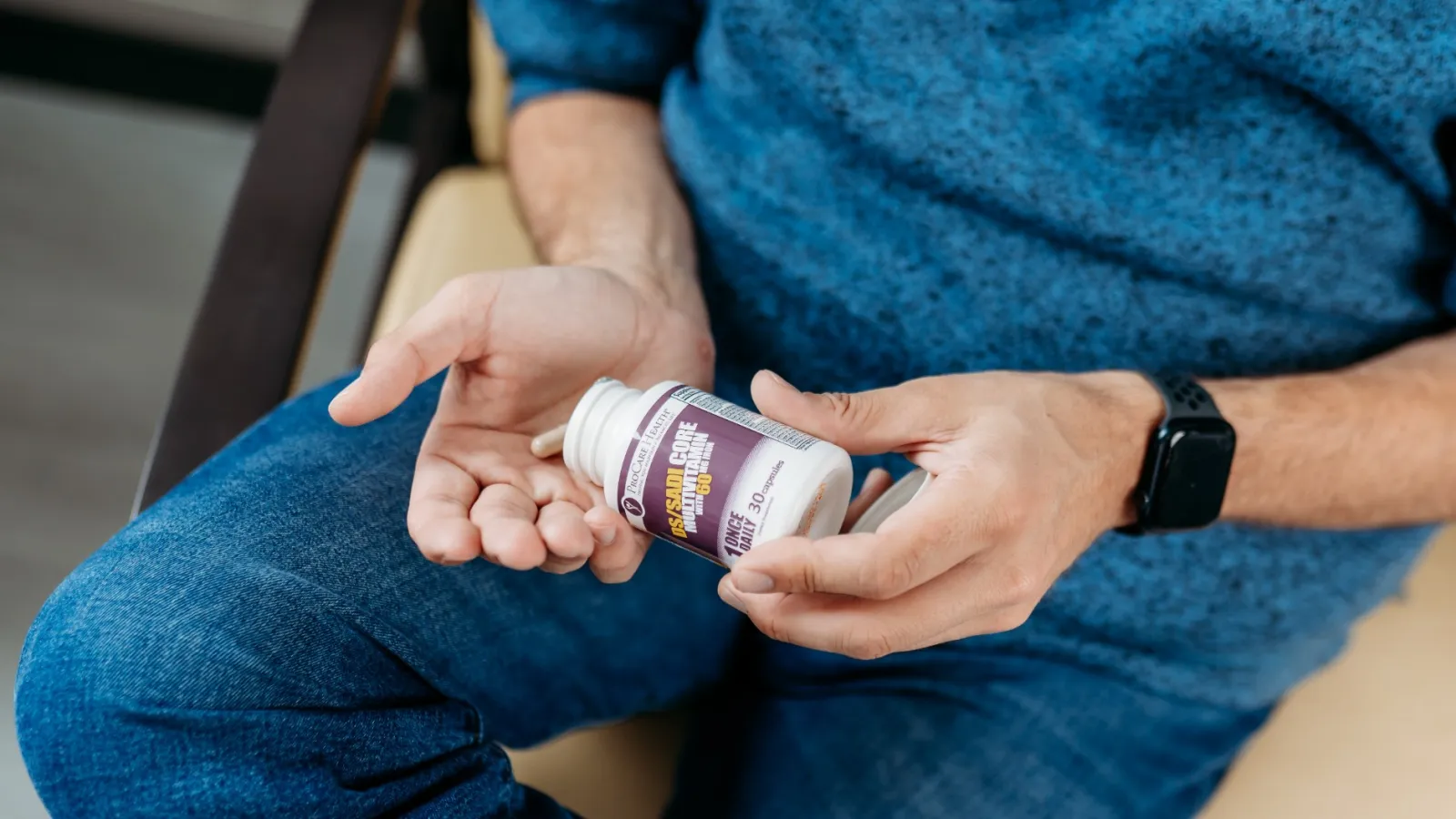 Man wearing blue sweater holding a bottle of capsules and placing pills in his hand while sitting on a chair.