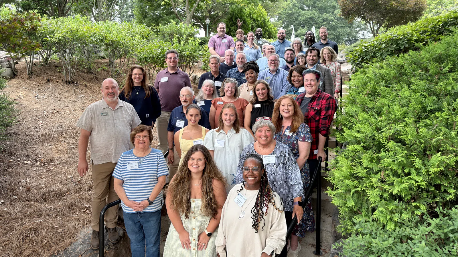 Group photo of diverse adults smiling on outdoor stairs surrounded by trees and greenery on a sunny day