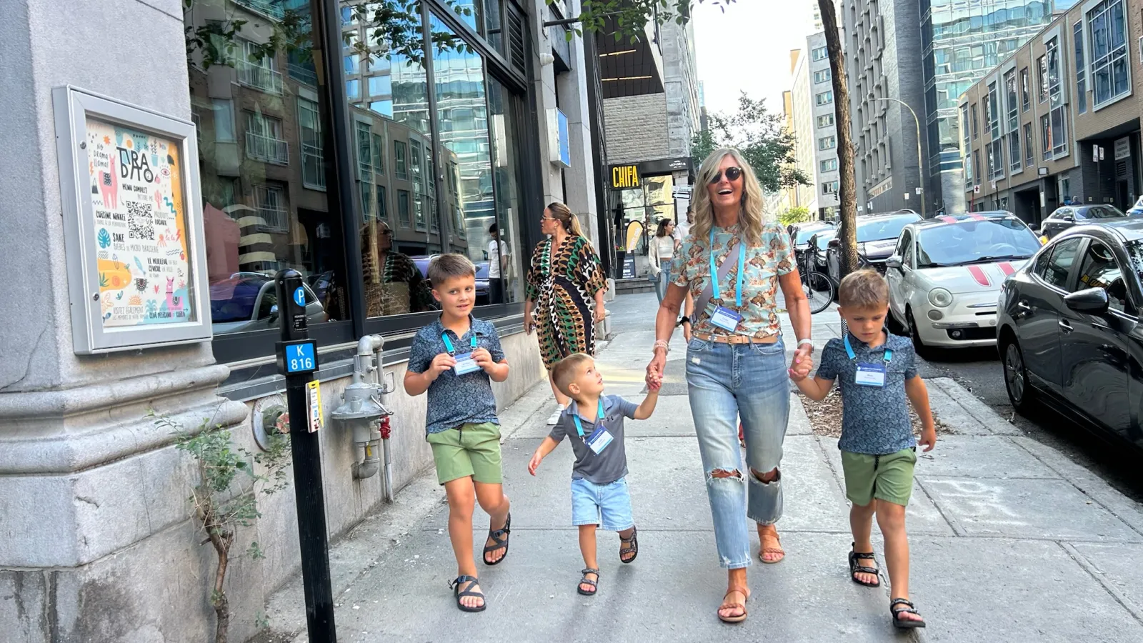 Smiling woman walking hand-in-hand with three young boys on a sunny city sidewalk with tall buildings.