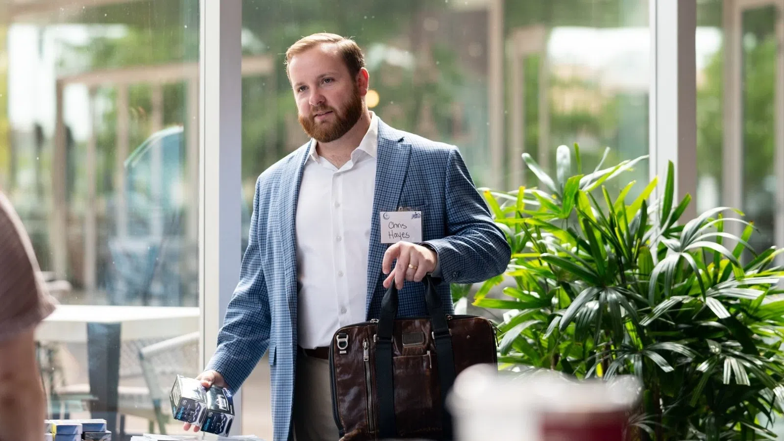 Man in blue blazer handing out name badges at a conference registration table indoors with plants and glass walls.