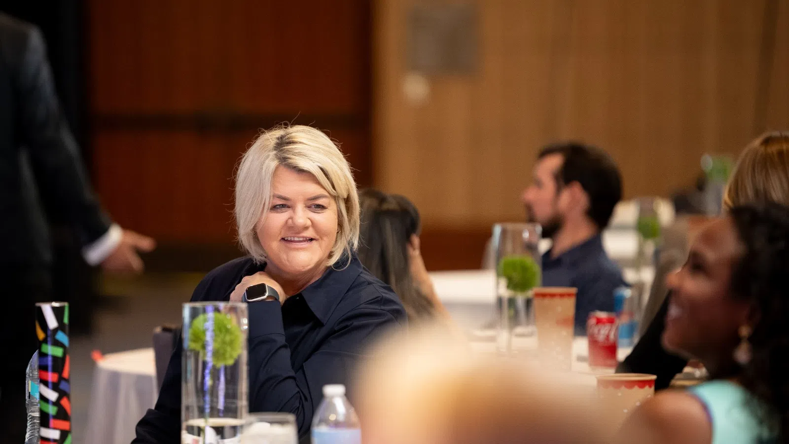 Smiling woman seated at a conference table with attendees and presenter in a professional setting.