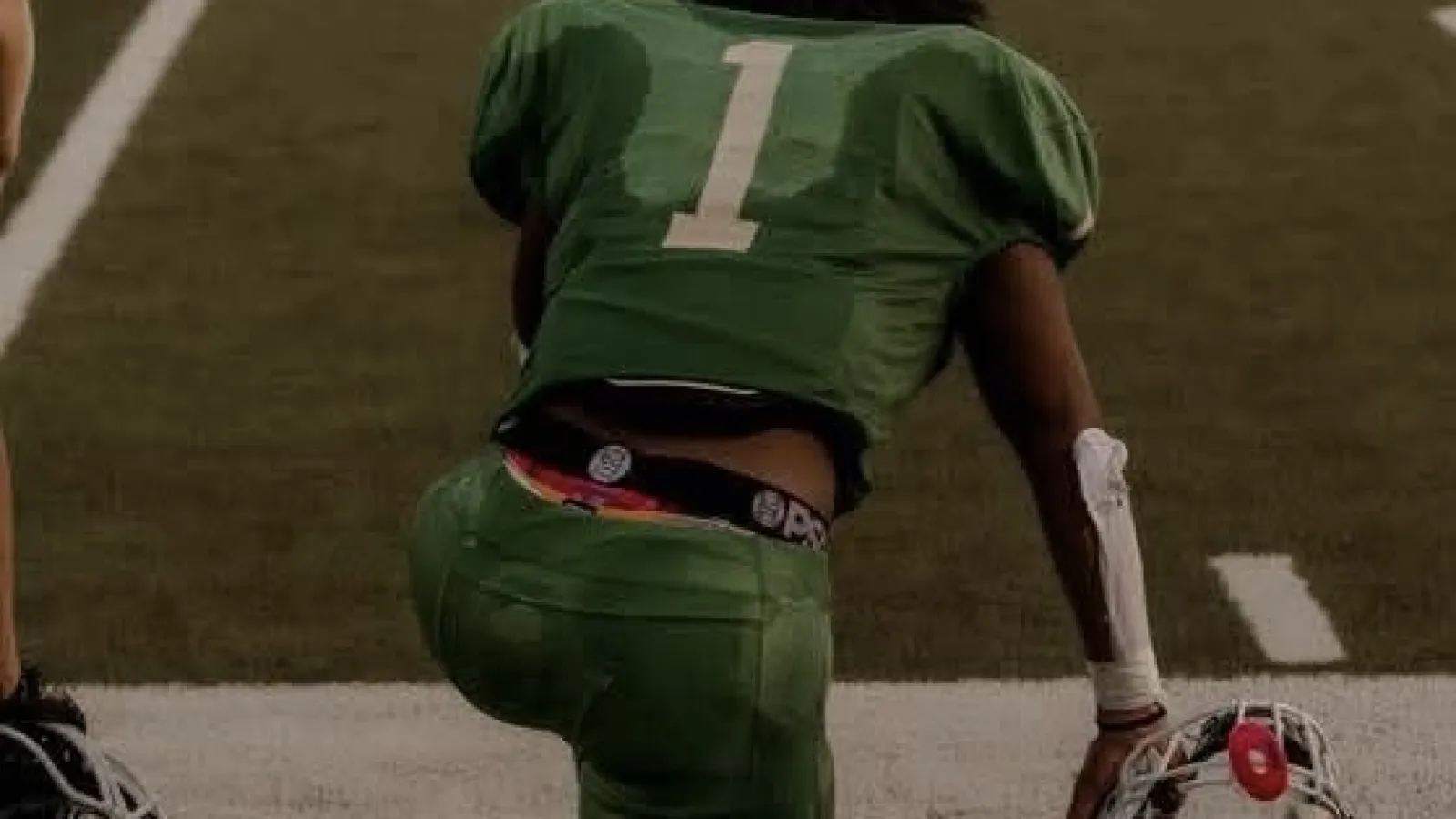 Football player wearing green jersey number 1 kneeling on field holding white helmet with red mouth guard.