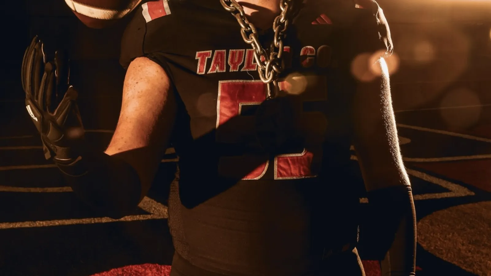 Football player in black Taylor County jersey tossing ball with chain necklace in dramatic lighting.