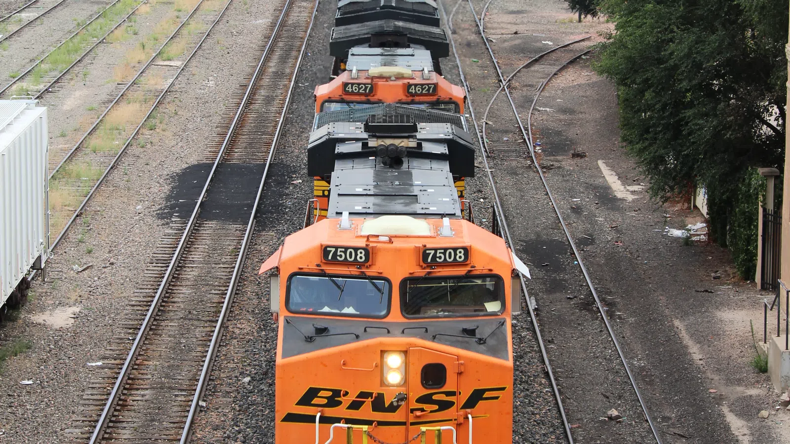 BNSF freight train with orange locomotives traveling on railway tracks near industrial buildings and mountains.