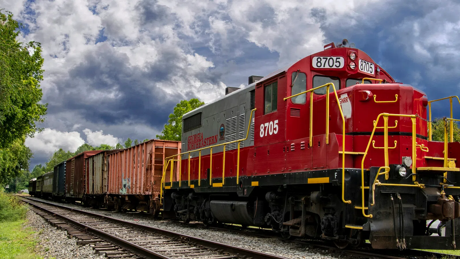 Red and gray diesel locomotive pulling freight cars on tracks under a dramatic cloudy sky.