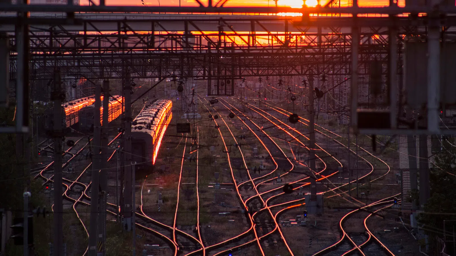 Train tracks reflecting sunset light with a stationary train and overhead electrical wires in an urban area.