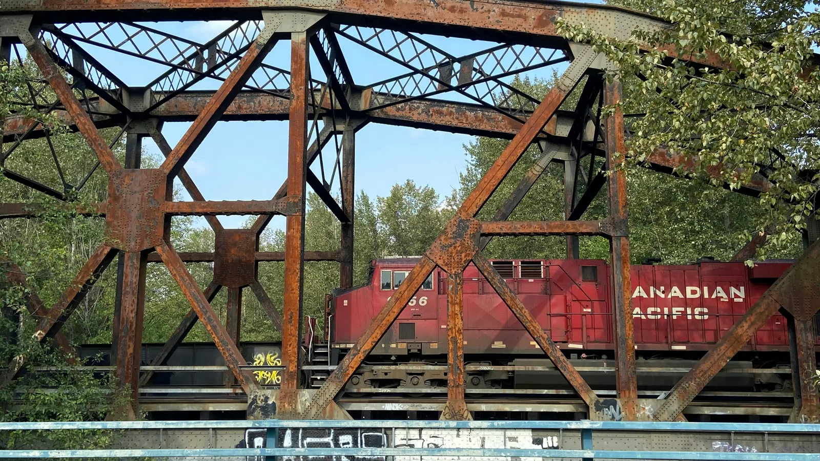 Rusty metal railway bridge with a red Canadian Pacific train crossing surrounded by trees under a clear sky.