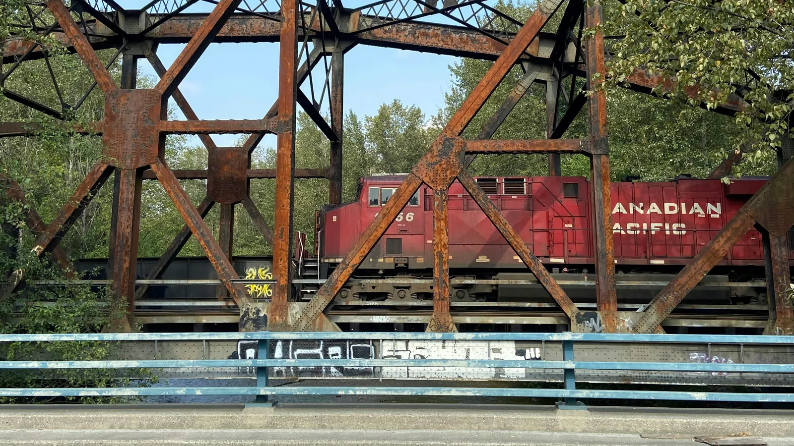 Rusty metal railway bridge with a red Canadian Pacific train crossing surrounded by trees under a clear sky.