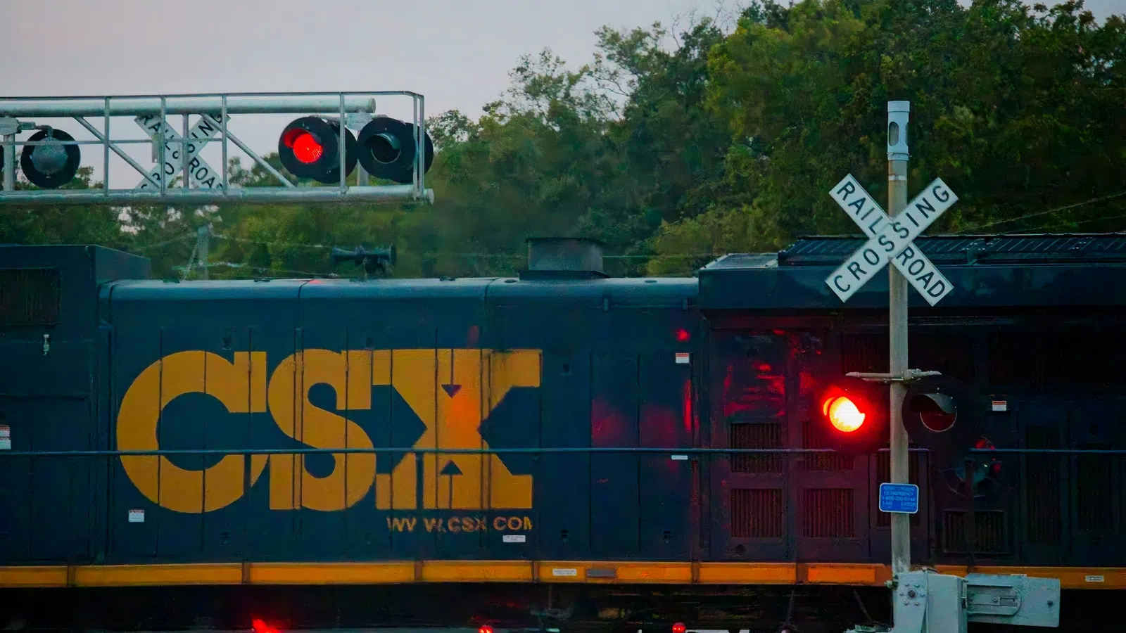 CSX freight train engine passing a railroad crossing with red signal lights and crossing barriers.