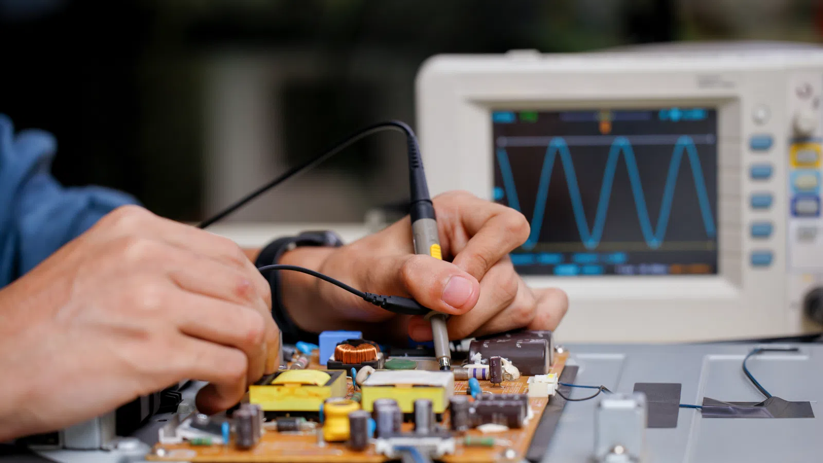Hands testing an electronic circuit board using probes with an oscilloscope showing waveform in the background
