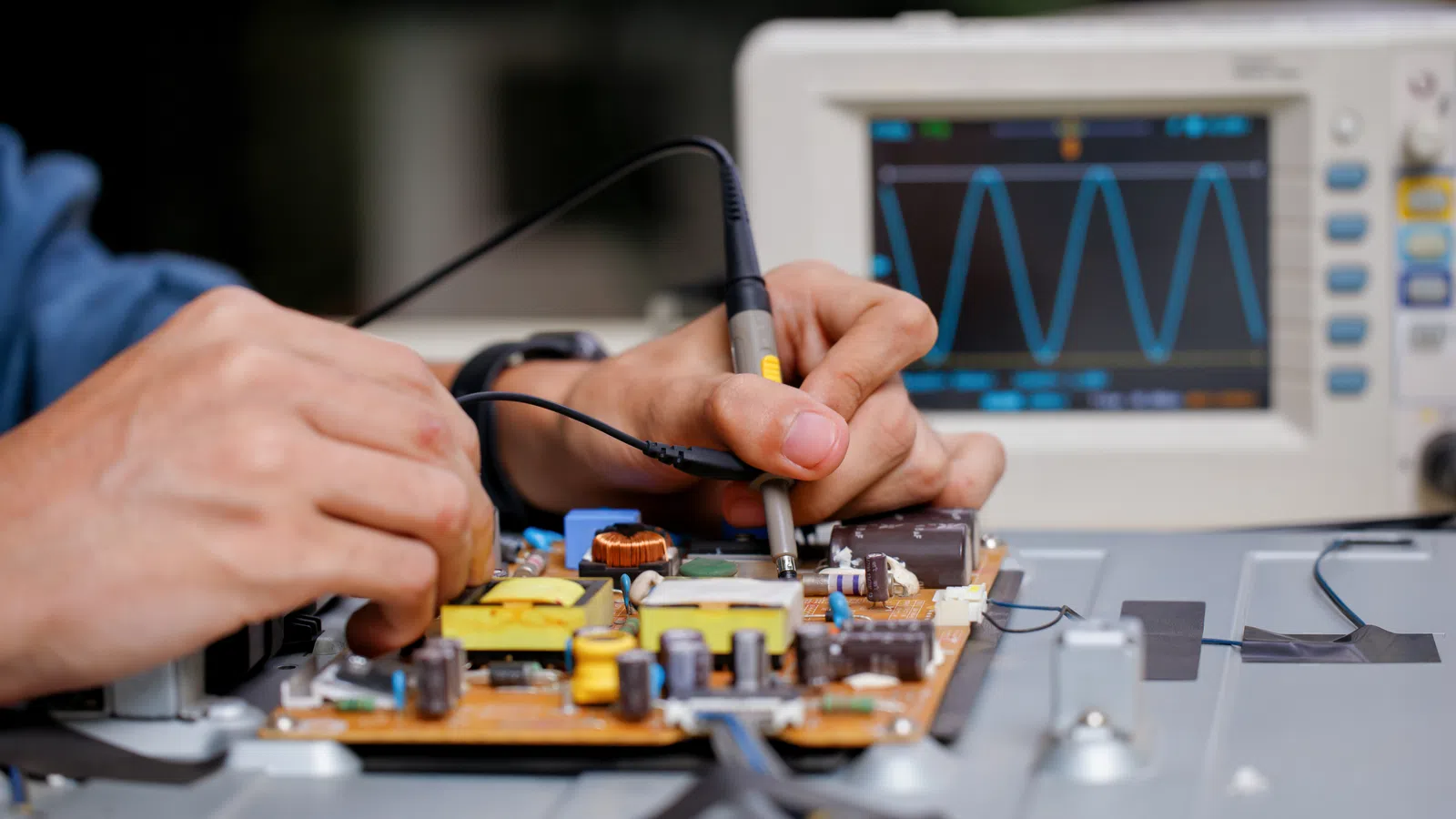 Close-up of hands testing electronic circuit board with oscilloscope displaying wave signal in background