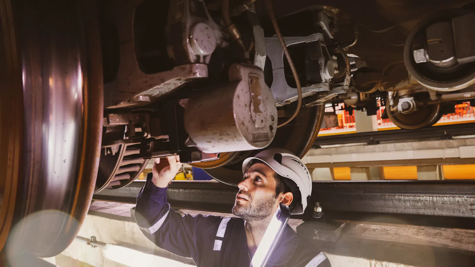 Engineer wearing safety helmet inspecting train undercarriage with flashlight in maintenance workshop.