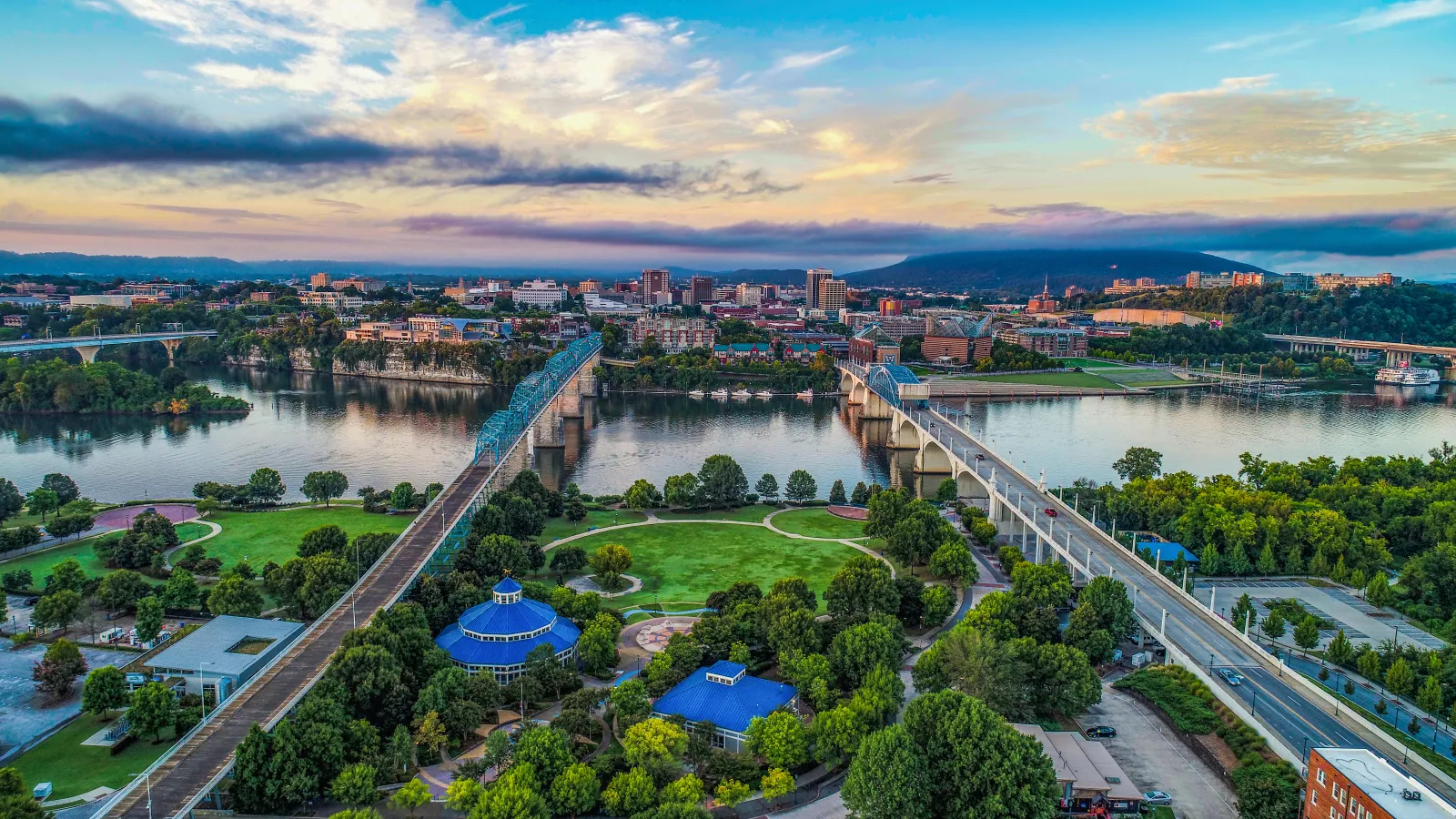 Aerial view of city with twin bridges over river, green park with blue-roofed buildings, and cloudy sky at sunset.