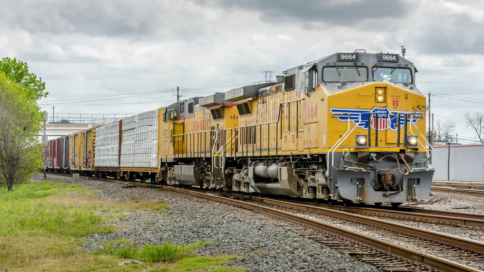 Yellow Union Pacific freight train moving on railroad tracks under cloudy sky near green foliage.