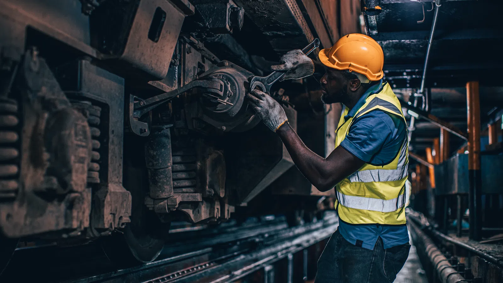 Worker wearing safety gear inspecting mechanical parts underneath a train in a dimly lit maintenance tunnel