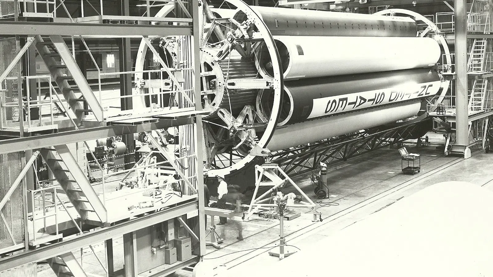 Large rocket stage under assembly inside industrial facility with scaffolding and workers.