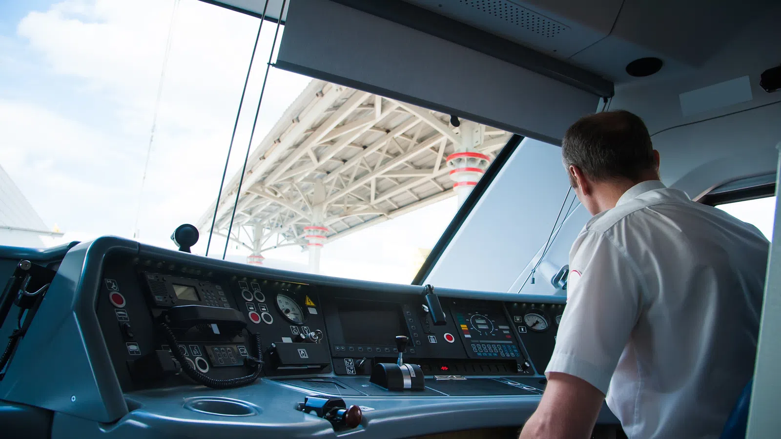 Train operator inside modern locomotive cab monitoring control panels under station roof.