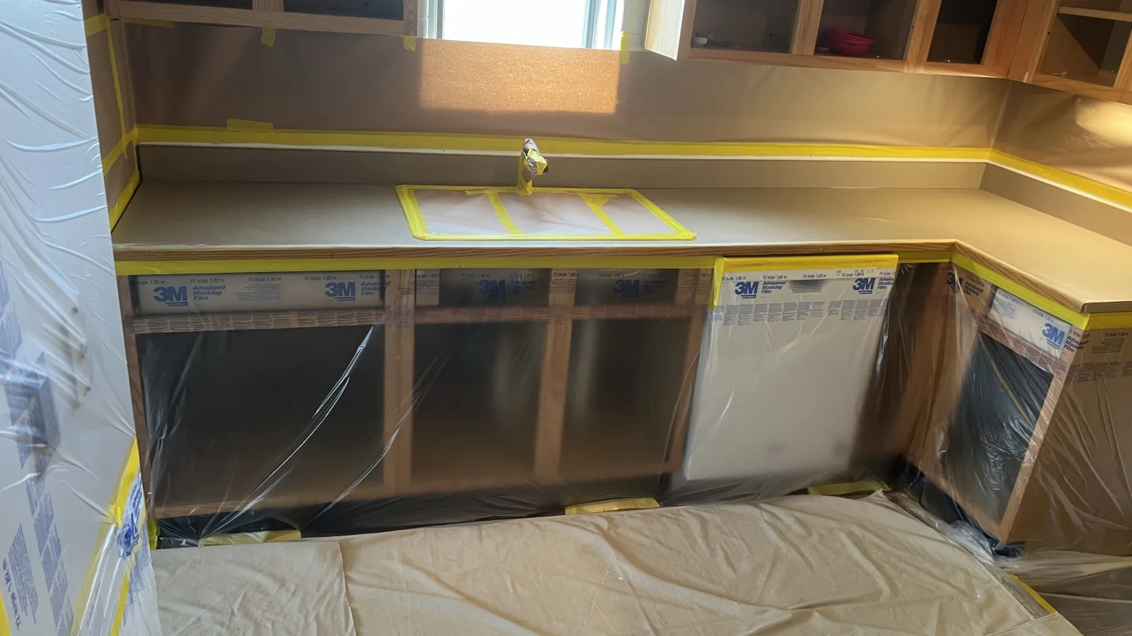 Kitchen cabinets and countertops protected with plastic and tape during renovation work under natural light.