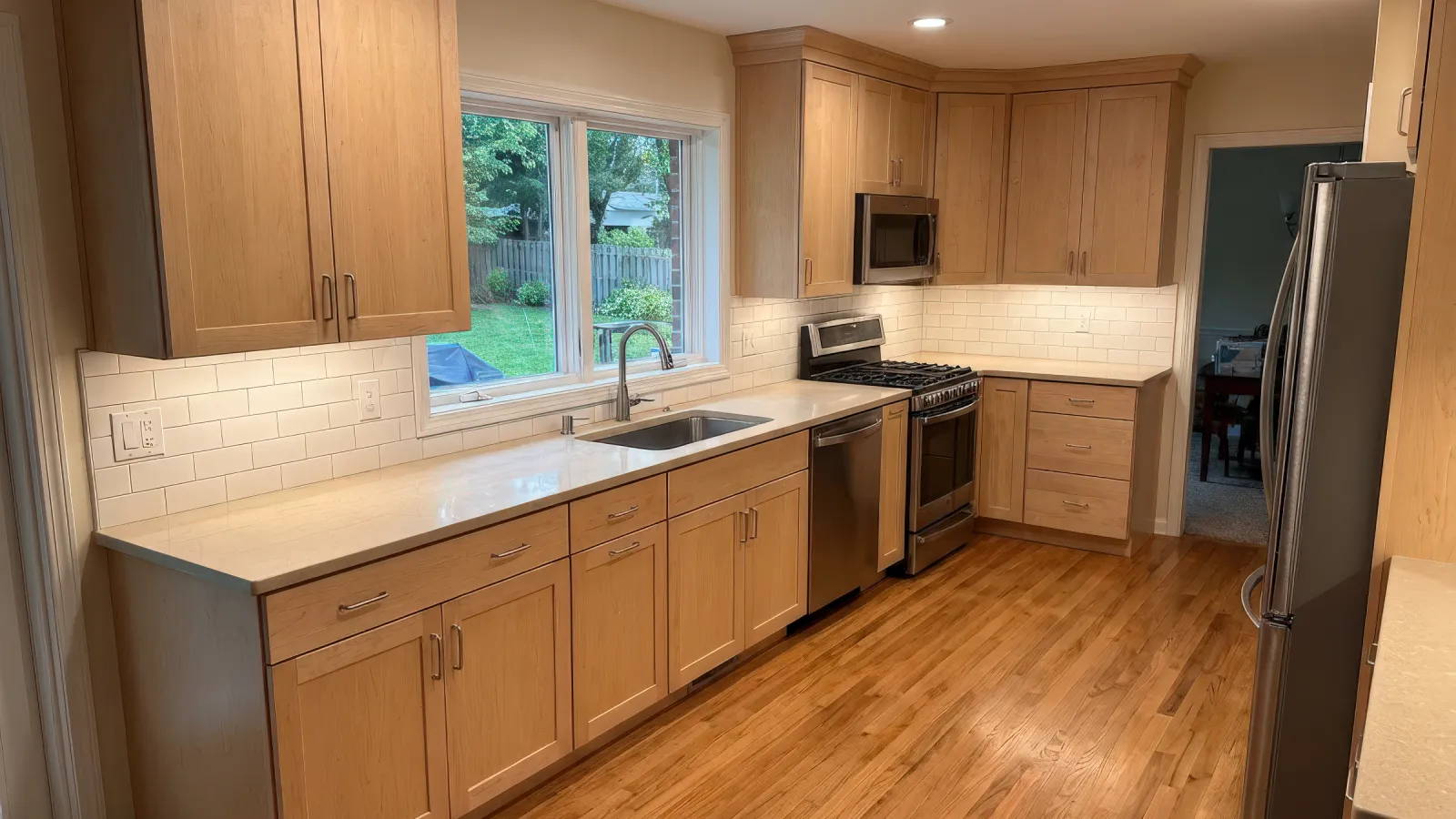 Modern kitchen with light wood cabinets, white countertops, stainless steel appliances, and hardwood flooring