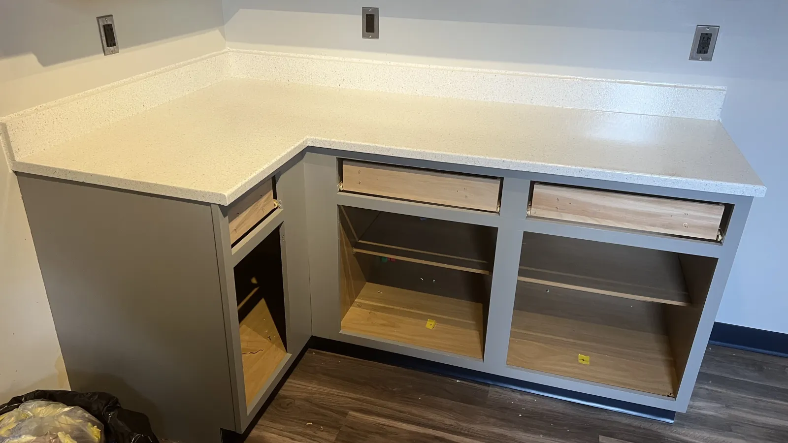 Newly installed L-shaped kitchen cabinets with white quartz countertop and empty wooden shelves beneath.