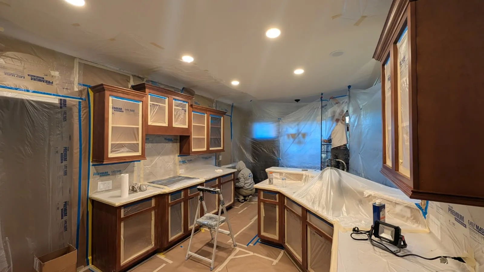Kitchen renovation in progress with covered cabinets, countertops, and workers preparing surfaces under recessed lighting.