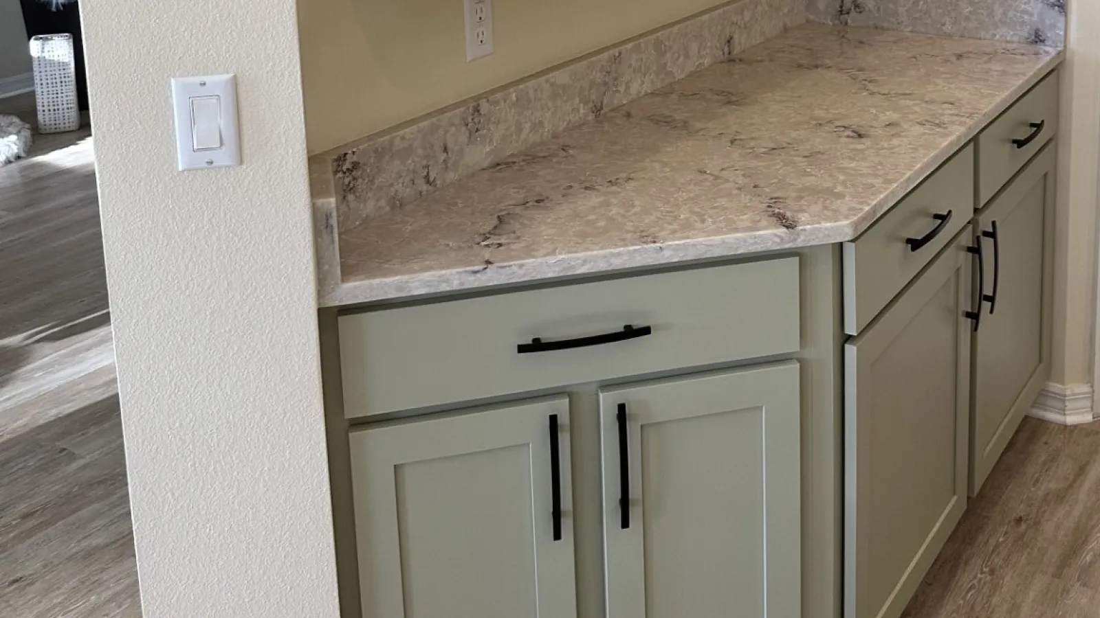 Kitchen corner with light gray cabinets, black handles, marble countertop, and wood-look flooring.