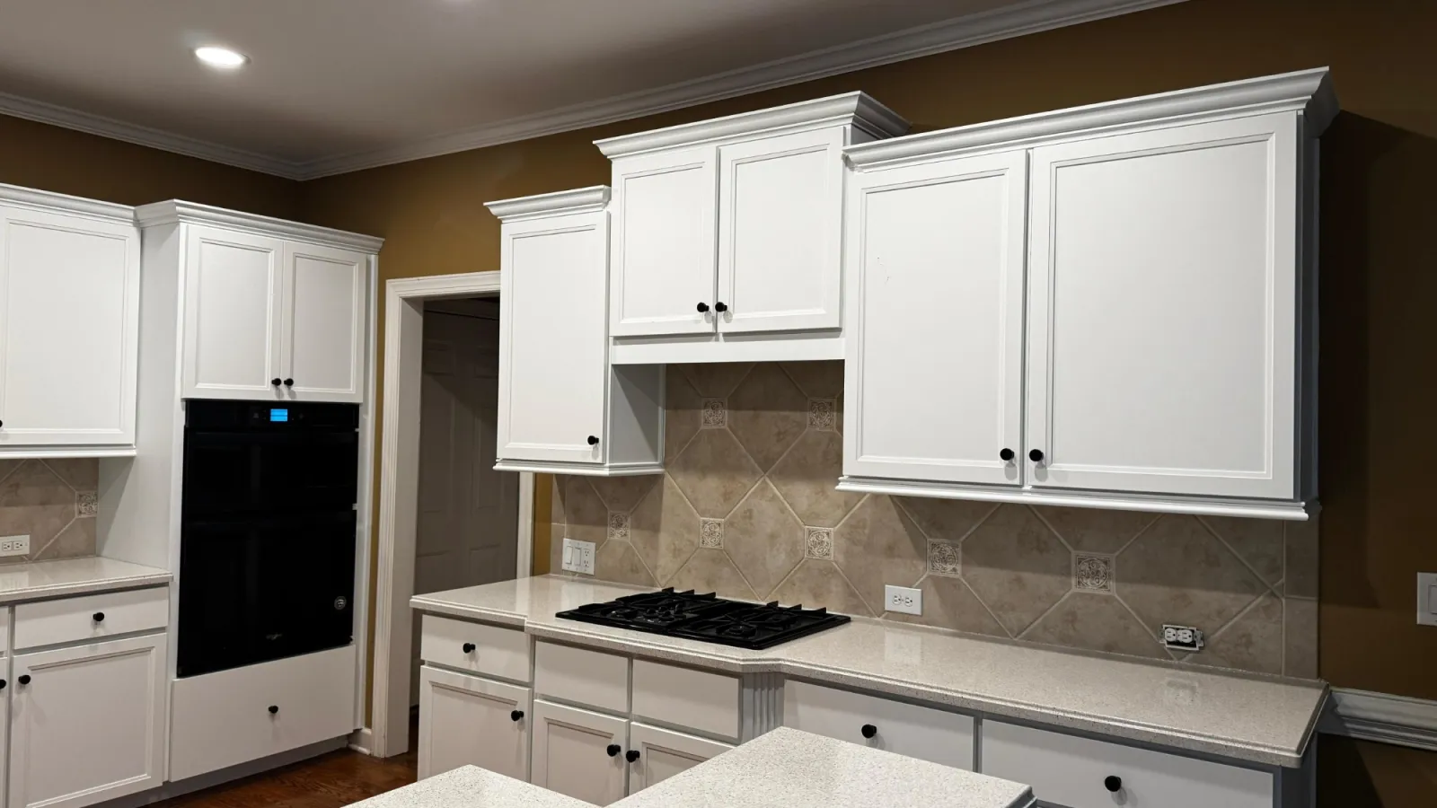 Bright modern kitchen with white cabinets, beige tiled backsplash, built-in black oven, and gas cooktop under recessed ceiling lights