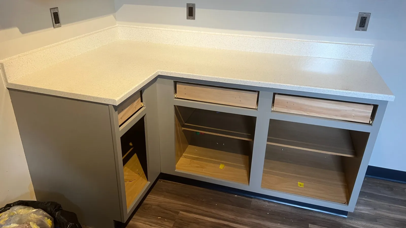 Newly installed L-shaped kitchen cabinet with white countertop and wooden floor in progress with drawers missing.