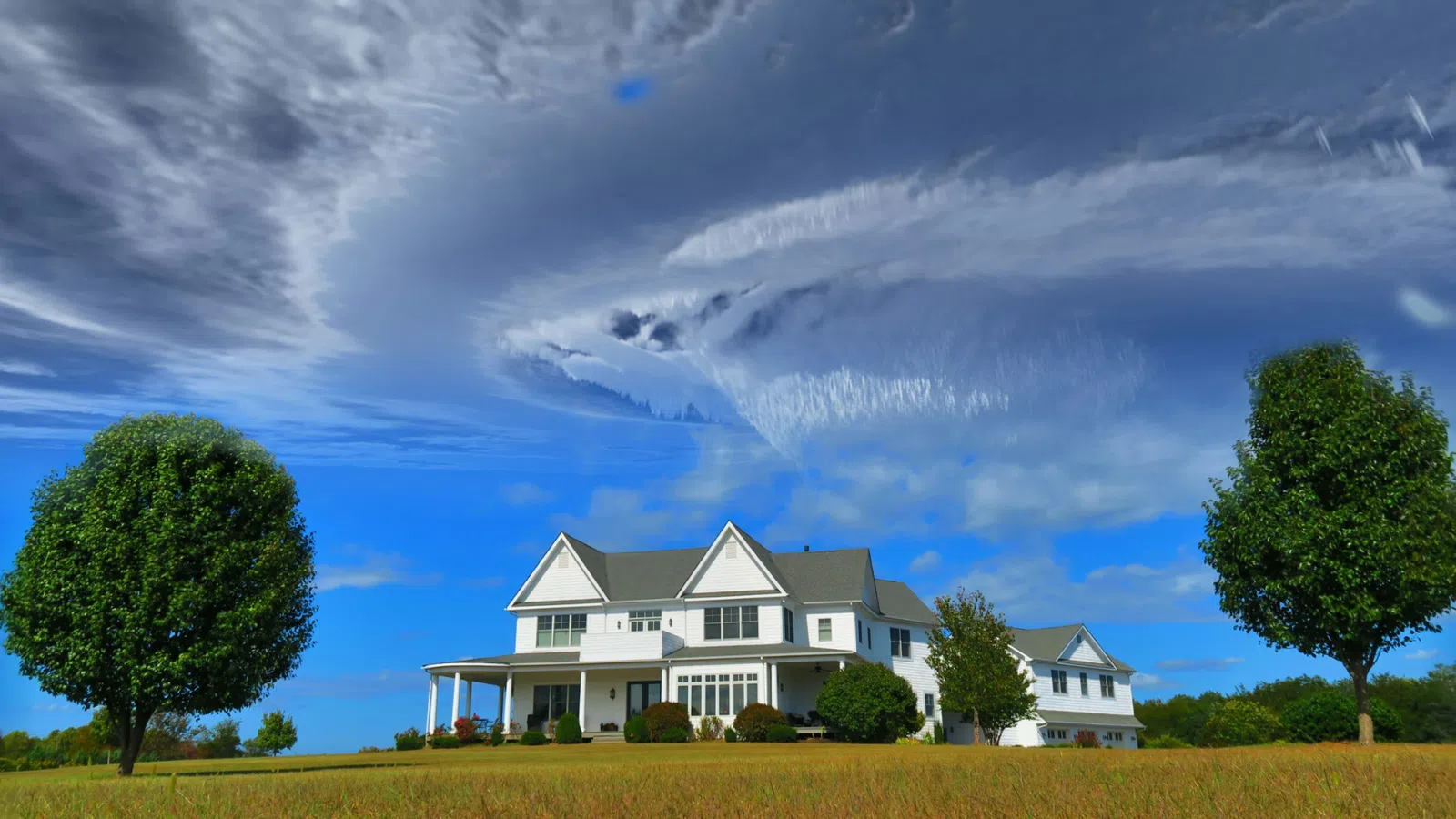 Large white farmhouse with wraparound porch between two trees under dramatic swirling clouds and blue sky