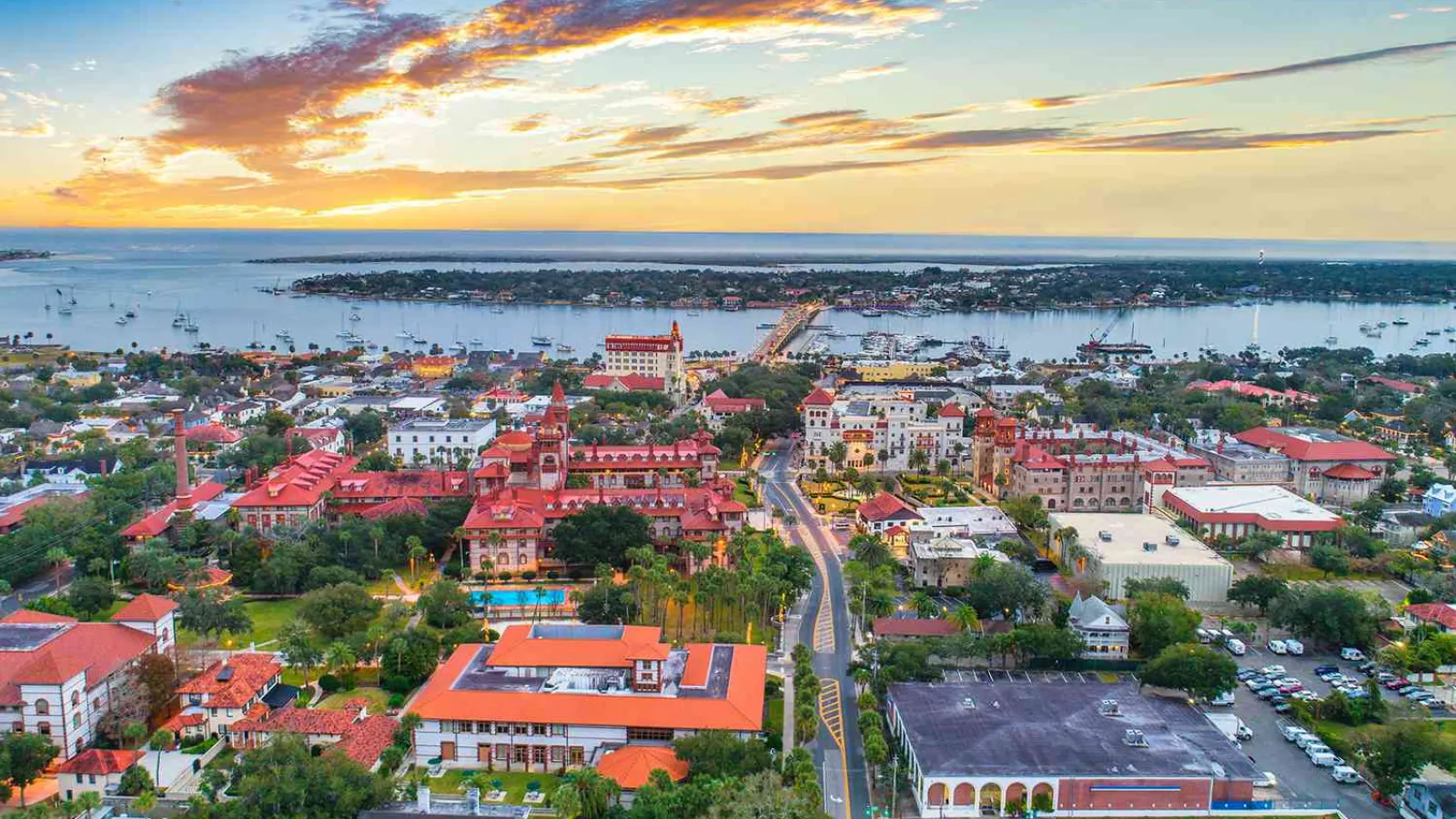 Aerial view of a coastal town with historic buildings, red roofs, a bridge, and a vibrant sunset sky.