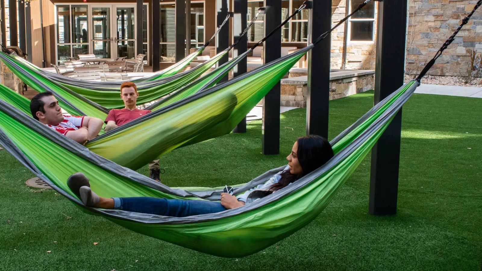 Three people relax in green hammocks in a vibrant outdoor space with modern architecture.