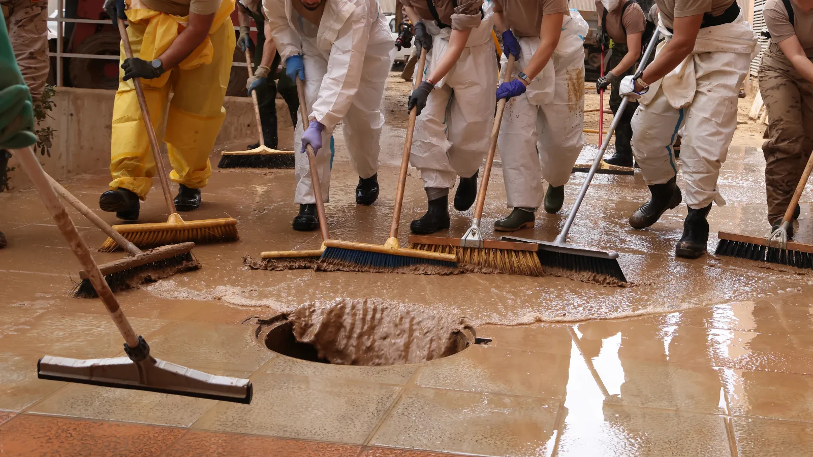 Group of people cleaning muddy water off floor near large drain wearing protective gear and gloves