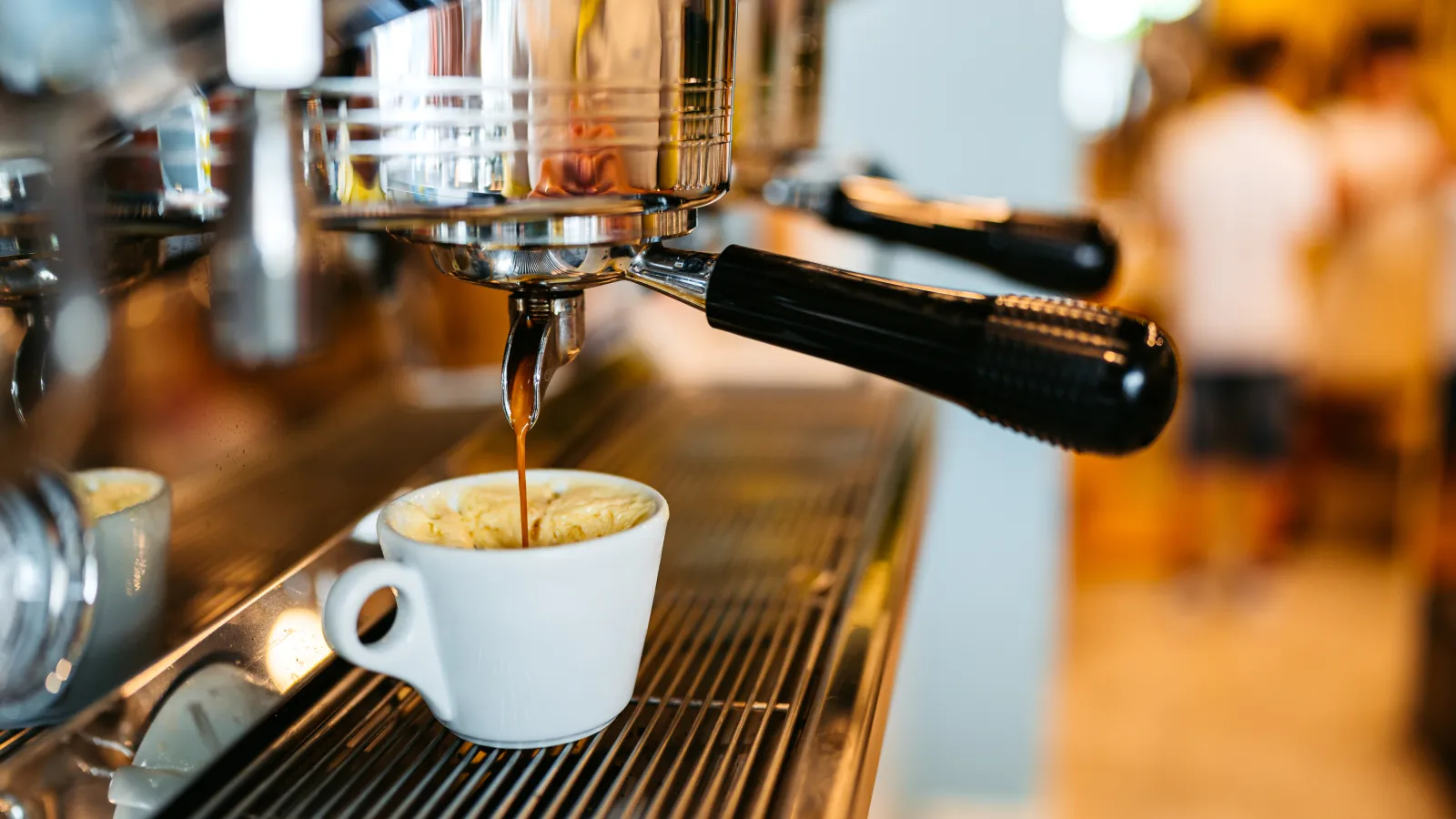 Espresso pouring from machine spout into white cup on coffee shop counter with blurred background.