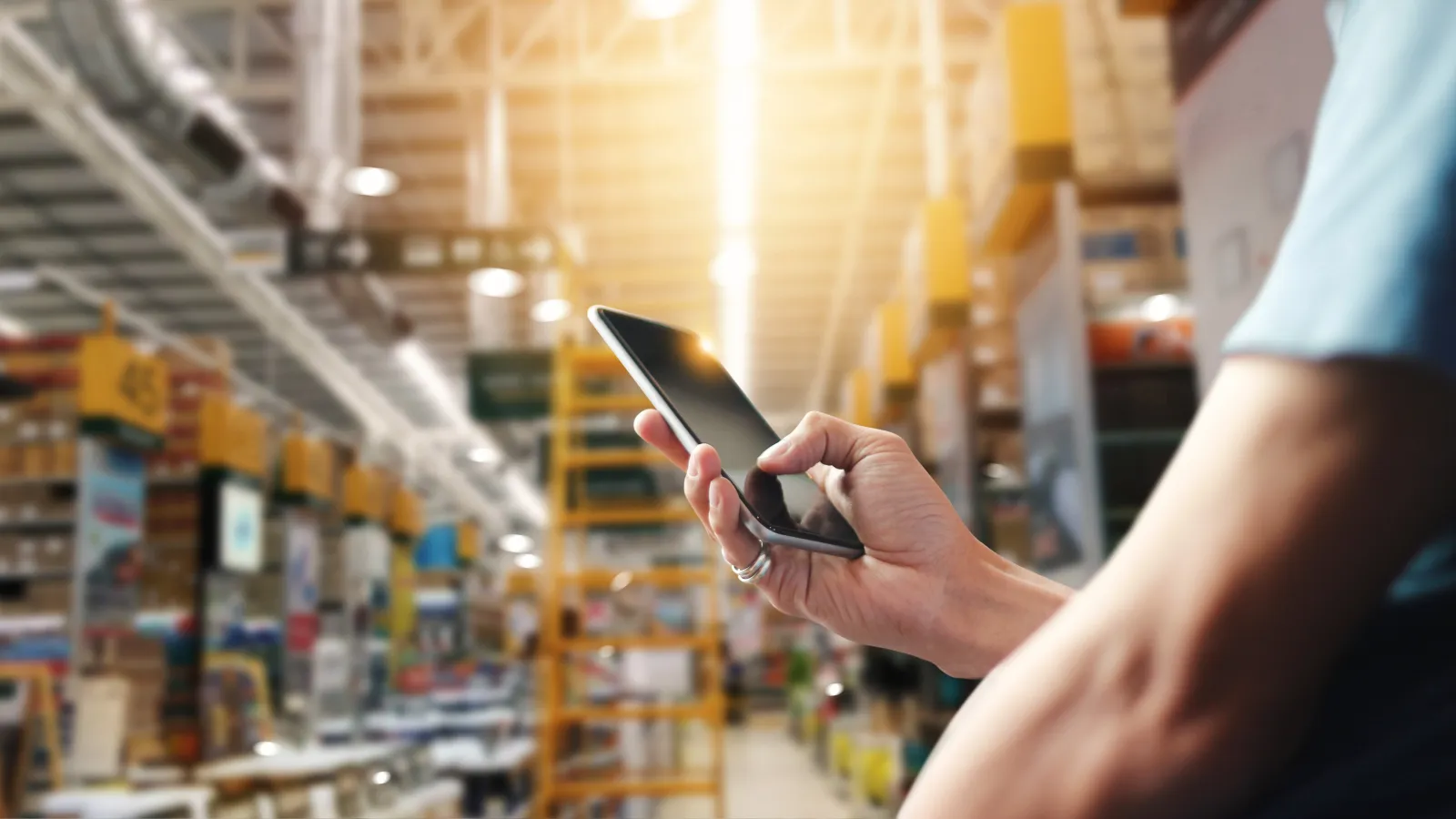 Person using smartphone inside a large, well-lit warehouse with tall shelves and products.