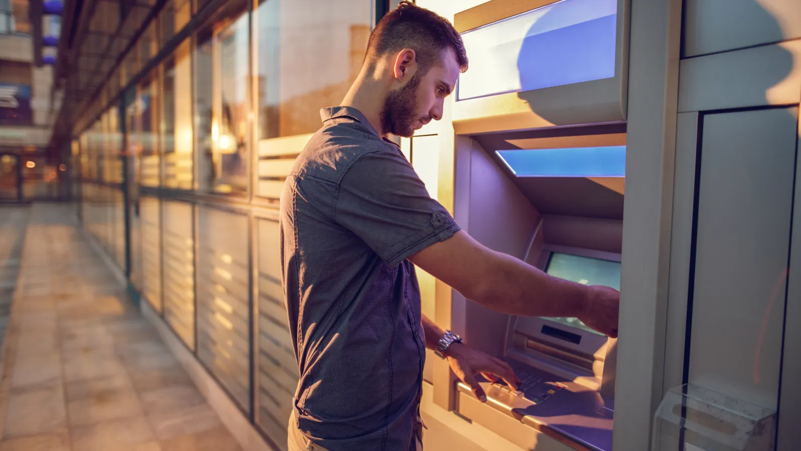 Man using an ATM outside a glass building during sunset, entering information on the keypad.