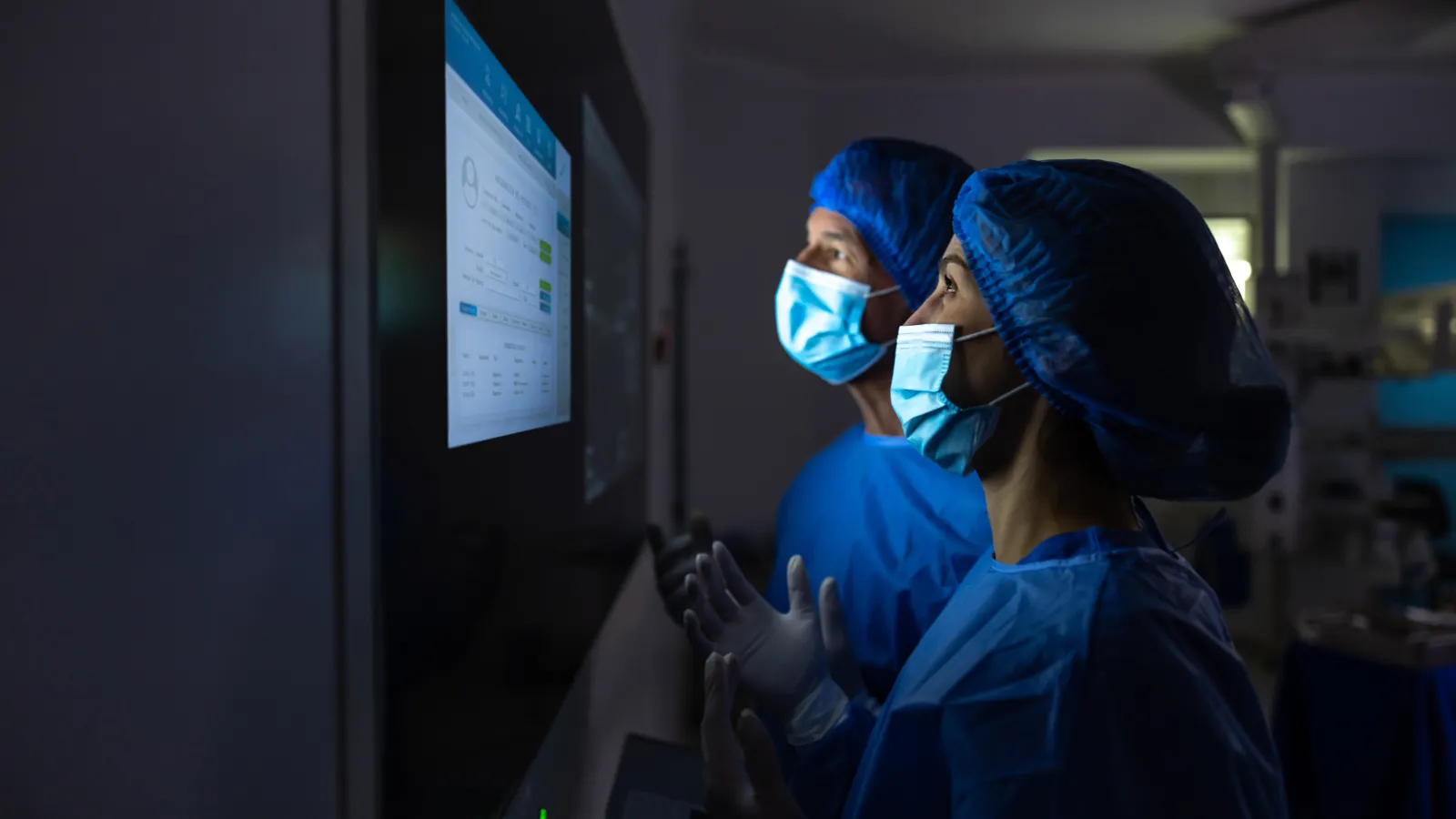 Two healthcare workers in masks and scrubs analyze medical data on a screen in a dimly lit room.