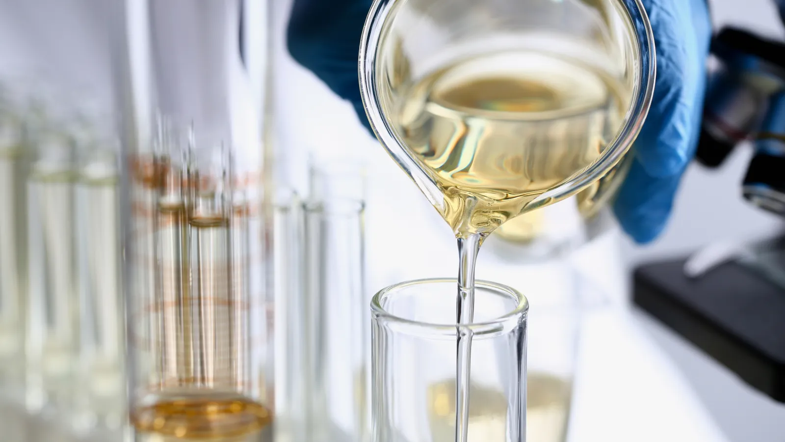 Scientist wearing glove pouring clear liquid from flask into test tube in laboratory setting with microscope.