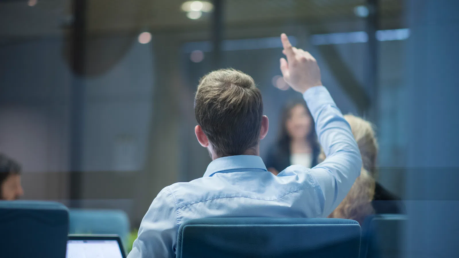 Man in office raises hand during meeting, colleagues and laptop visible in modern conference room.