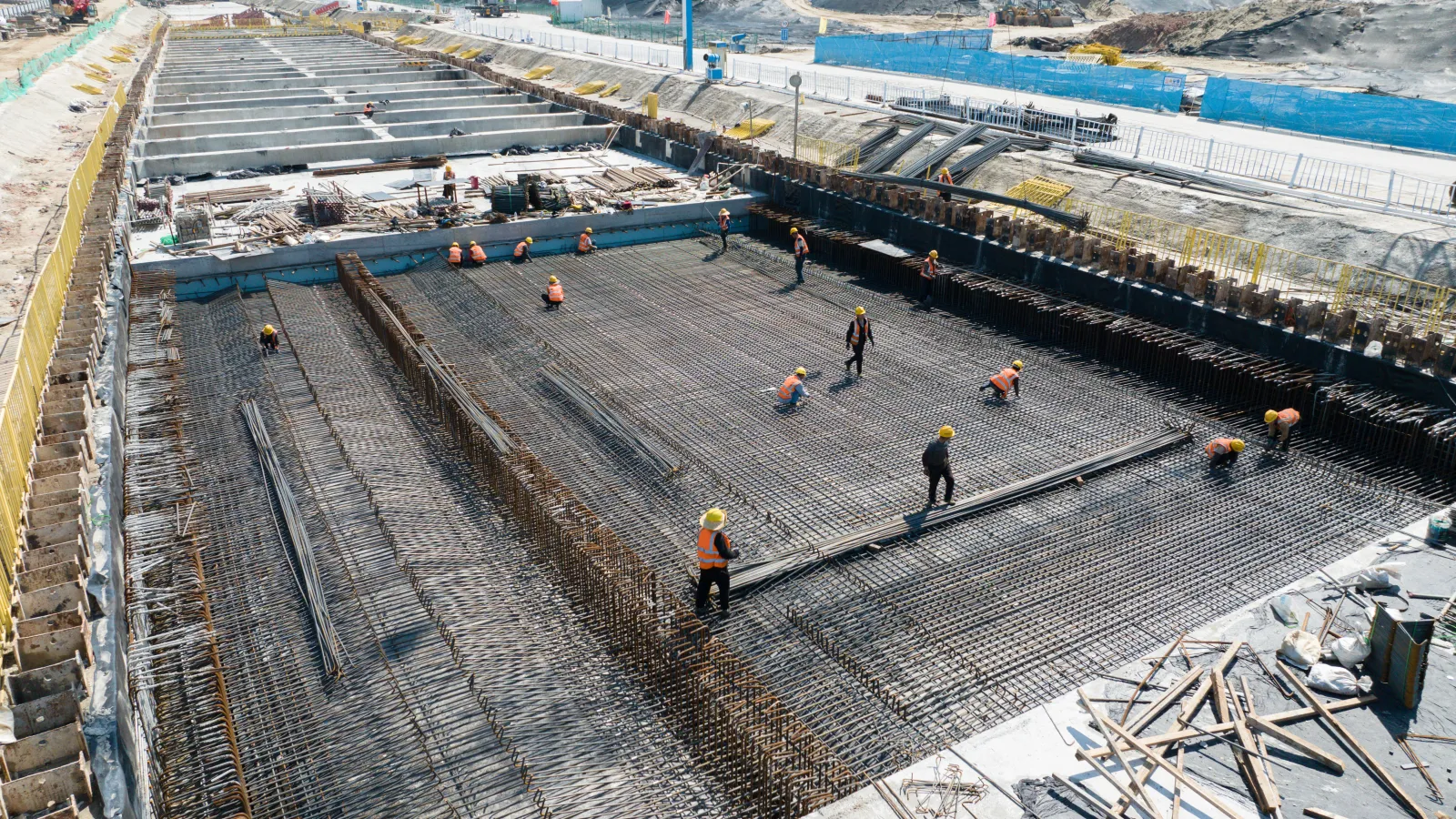 Construction workers setting steel rebar for large concrete foundation at an industrial site under clear sky