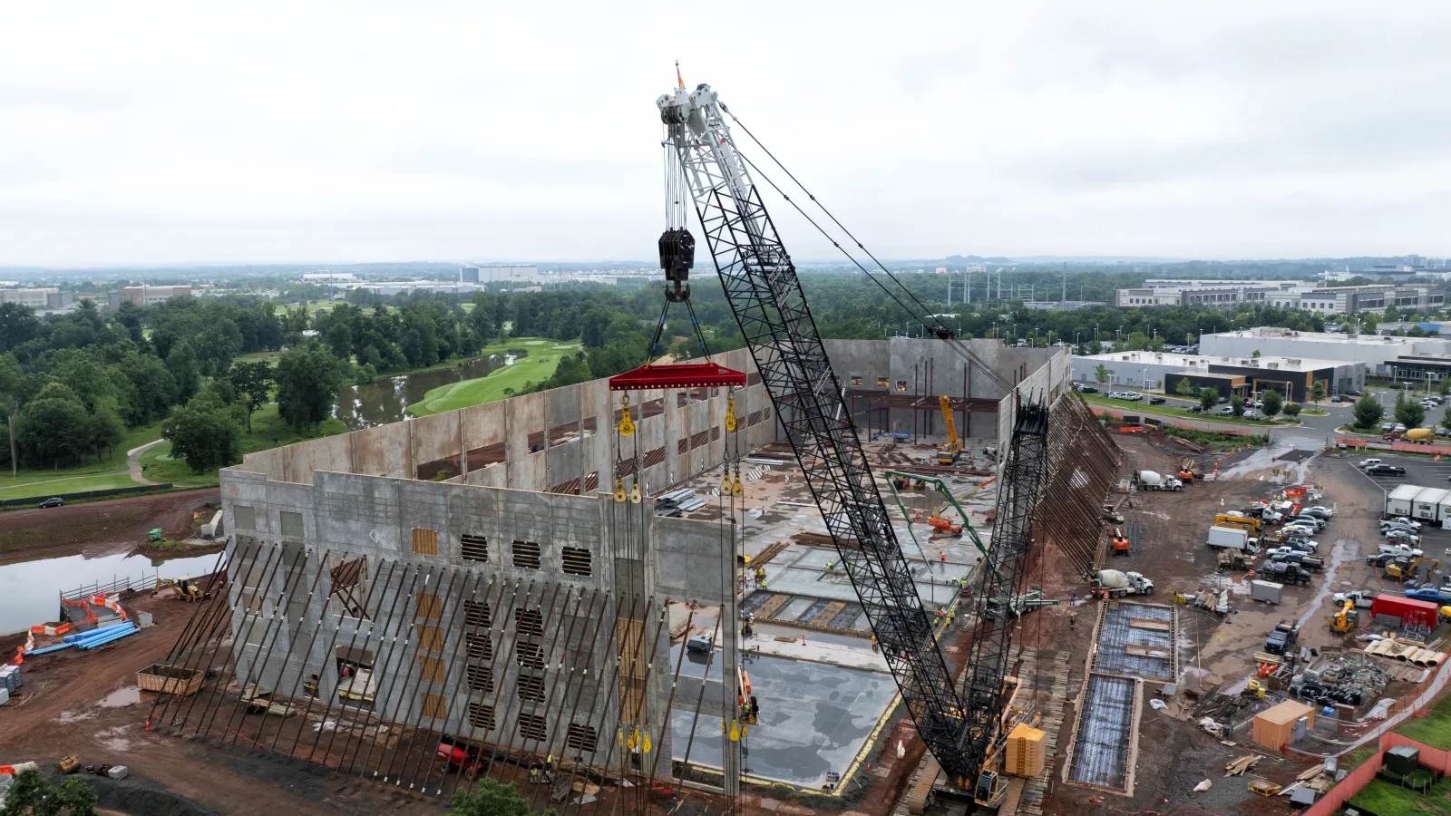 Large crane lifting concrete wall panels at a sprawling construction site with equipment and green surroundings.