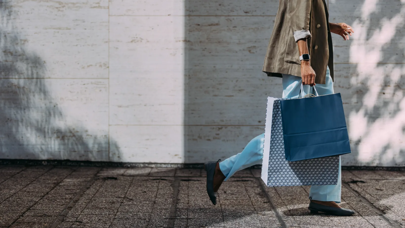 Person walking outdoors carrying blue and patterned shopping bags against a stone wall in daylight