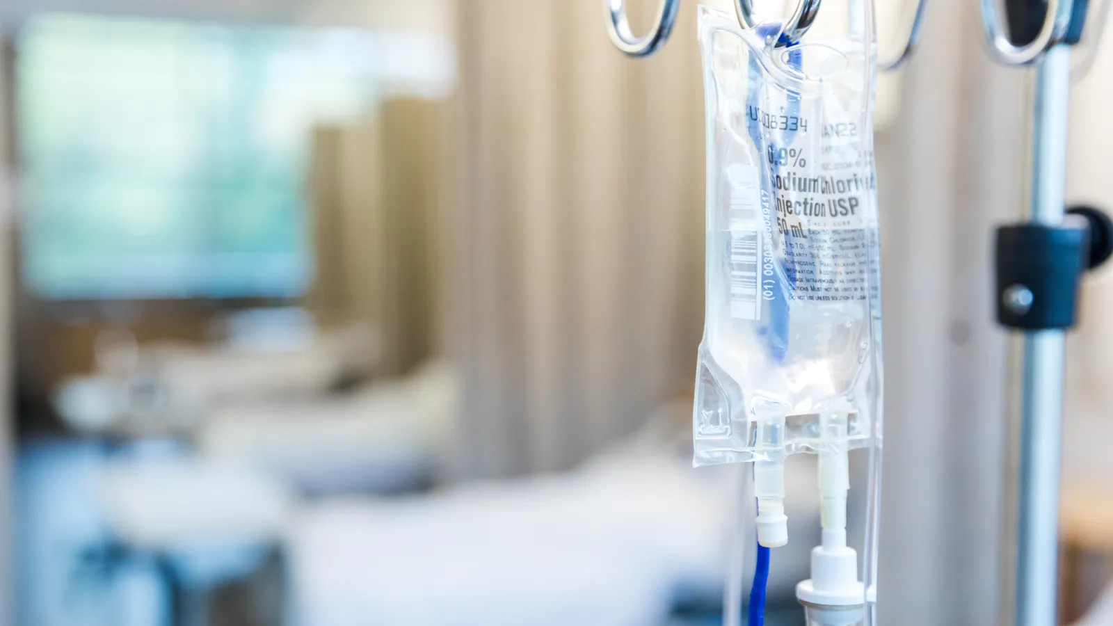 Close-up of an IV drip bag hanging on a metal stand in a hospital room with blurred beds in background