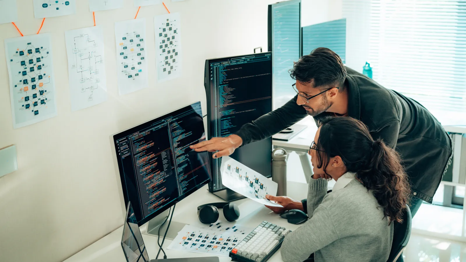 Two programmers collaborating on code at a desk with multiple screens and flowcharts on the wall.