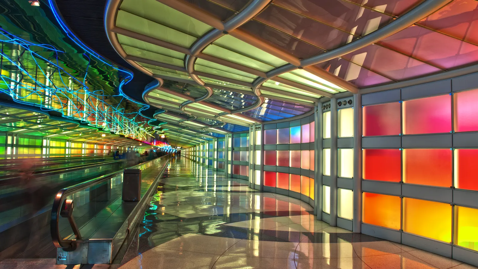 Colorful illuminated tunnel with curved ceiling and reflective floor, featuring vibrant gradient panels and moving walkway.