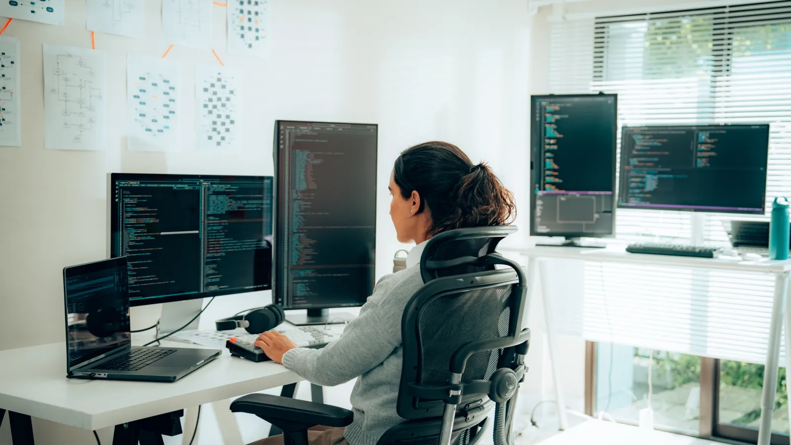 Woman coding at a modern desk with multiple monitors displaying programming languages and charts.