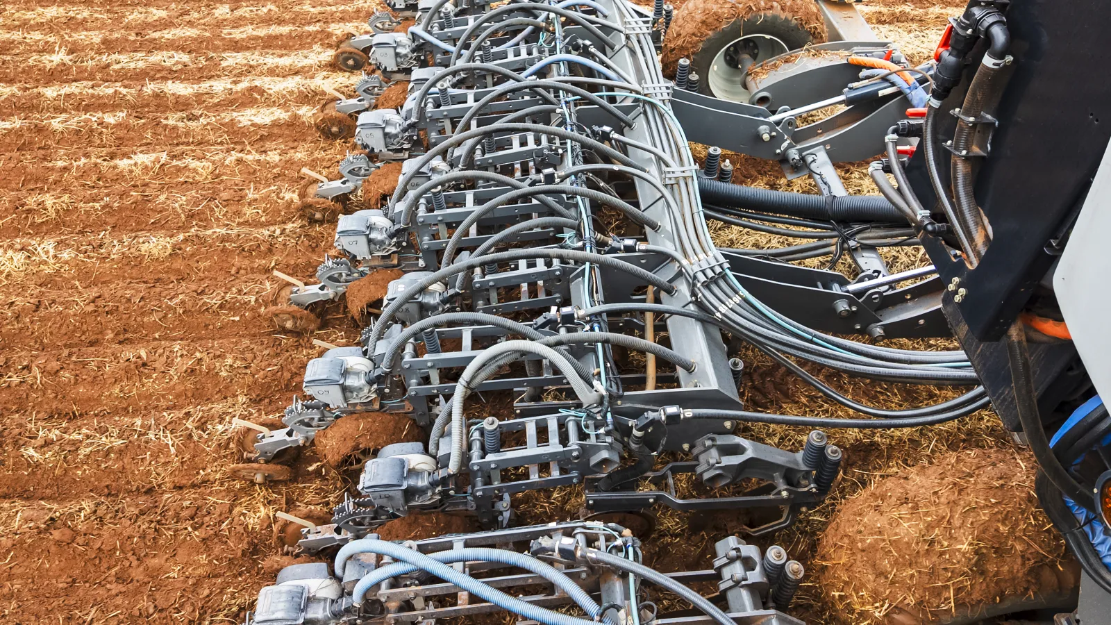 Modern agricultural machinery tilling dry soil in a field with multiple metal plows and hydraulic pipes.