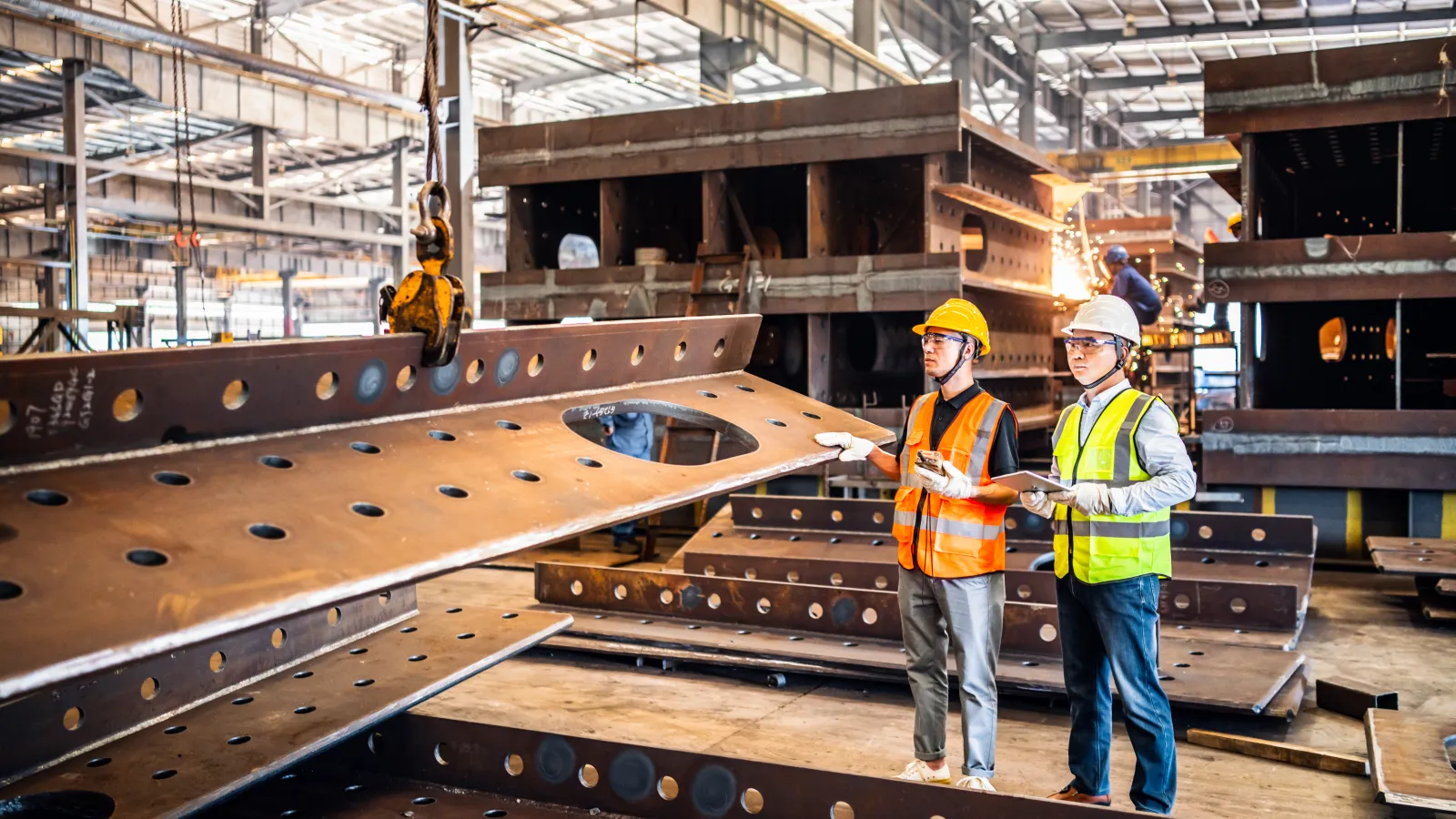 Two engineers in safety gear inspecting large steel beams in a spacious industrial factory setting.
